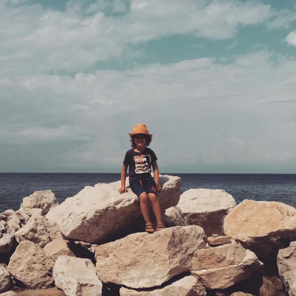 A child wearing a yellow hat sitting on coastal rocks by the sea under a clear sky. Vintage summer aesthetic.