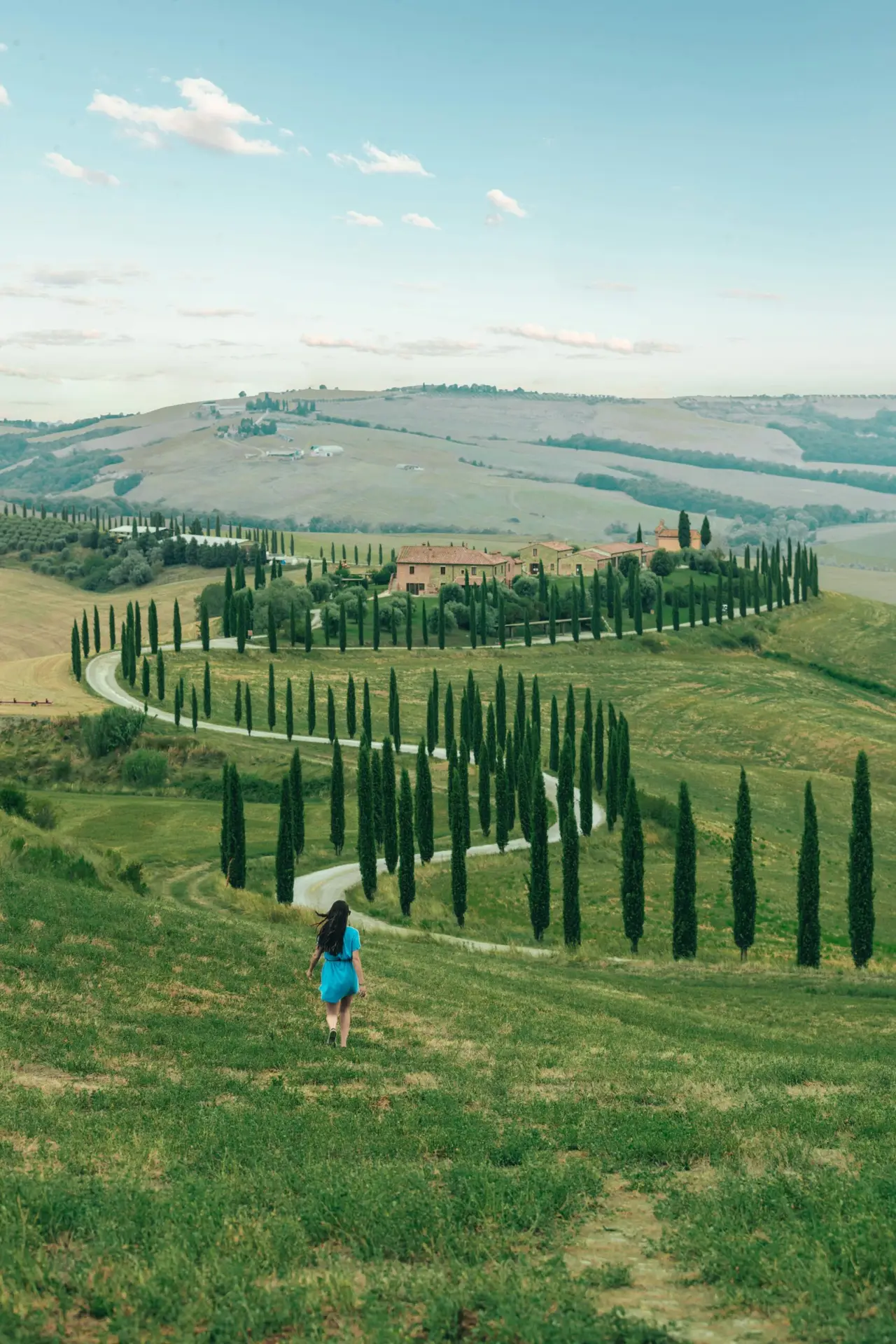 Serene view of a woman walking through picturesque Tuscan countryside with winding road and cypress trees.